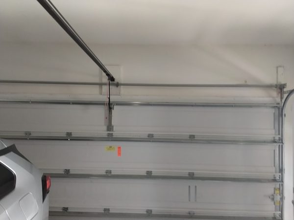 Closed white sectional garage door seen from inside a Chesapeake garage, with the horizontal spring tube running the full width above the top section and no exposed torsion springs, opener rail and red emergency release cord overhead, car parked at left