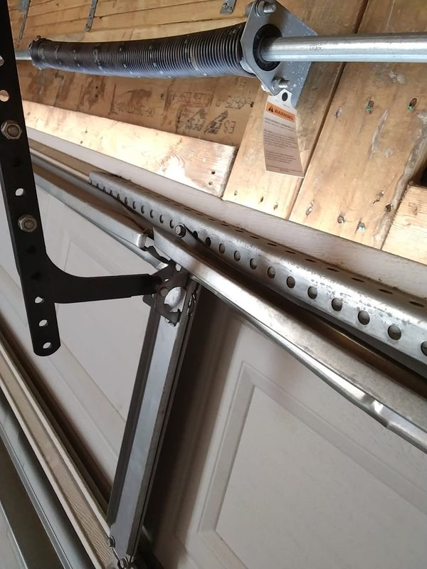 Bent and twisted galvanized reinforcement strut across the top section of a closed garage door, deformed where the opener arm bracket bolts on, with the torsion spring and shaft above, Norfolk Virginia
