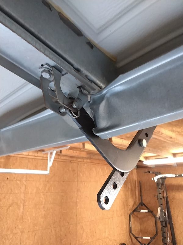 Close view looking up at the center of a garage door top section, showing the galvanized strut crushed and folded inward at the point where the opener arm bracket and cotter pin attach