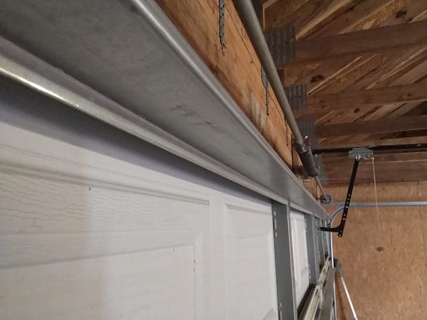 New straight galvanized 16 ft reinforcement strut installed across the top section of a closed garage door, seen from inside along the length of the door with the opener rail and torsion shaft above